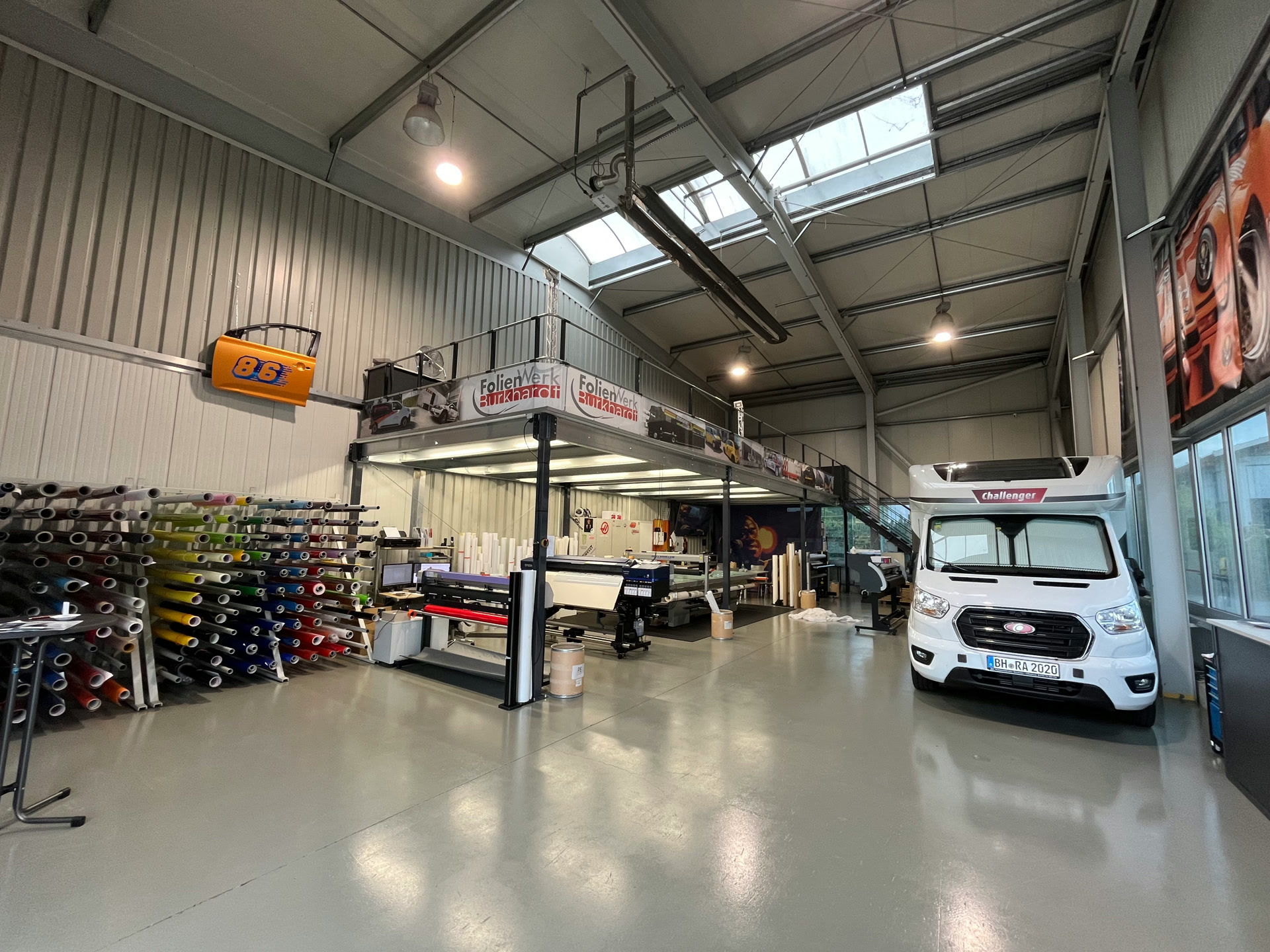 Die helle Werkstatthalle mit Folienrollen, Hebebühne und einem Transporter